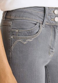 Vaqueros de mezclilla gris con costuras claras. Presentan un diseño ondulado en los bolsillos y dos botones decorativos en la cintura. Textura suave, corte ajustado.