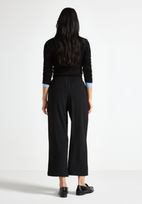 Pantalones negros de pierna ancha con cinturilla elástica, combinados con un top ajustado de manga larga negro y detalles azules en los puños. Se ven zapatos planos.