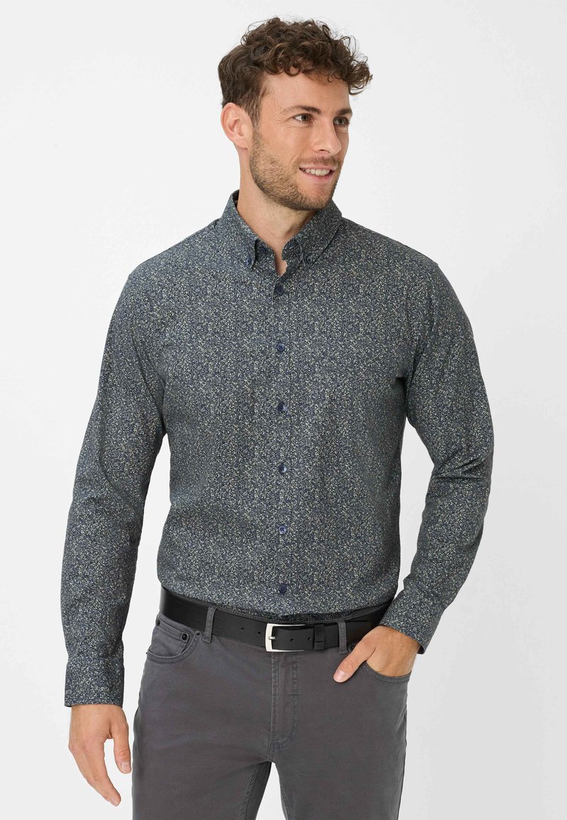 Mann mit lockigem Haar, der ein dunkelblümchenbedrucktes Hemd und graue Hosen trägt, lächelt und schaut zur Seite, mit einer Hand in der Tasche.