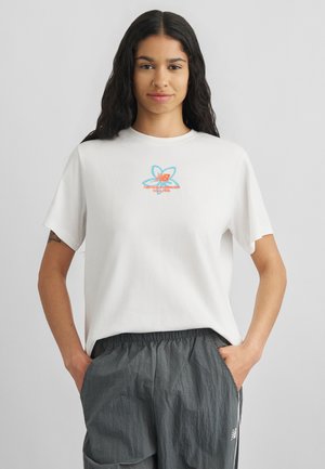 Young woman with long dark hair wearing a white t-shirt with a butterfly and New Balance logo, and loose gray pants with hands in pockets.