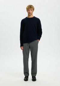Temno modra puhasta pletenina z okroglo izreznico, v paru s svetlo sivimi elegantnimi hlačami in črnimi čevlji. Model stoji z rokami v žepih.