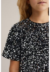 Musta t-paita, jossa on kiiltäviä hopea- ja pronssisequineja. Lyhyet hihat, pyöreä kaula-aukko, teksturoitu pinta tiheällä sequin-kuviolla.