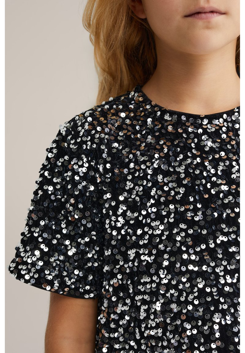 Musta t-paita, jossa on kiiltäviä hopea- ja pronssisequineja. Lyhyet hihat, pyöreä kaula-aukko, teksturoitu pinta tiheällä sequin-kuviolla.