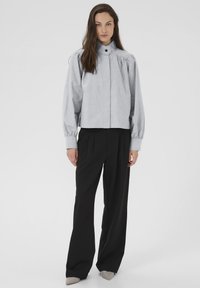 Kvinde står iført en lys grå knapbluse med rynkede skuldre og sorte bredbenede bukser, beige spidse sko, neutral baggrund.