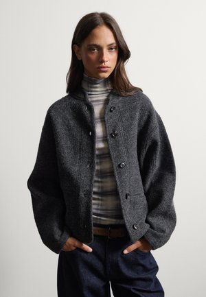 Joven mujer con cabello largo y oscuro que lleva una chaqueta de lana gris oscuro abotonada sobre un jersey de cuello alto a rayas y pantalones oscuros con las manos en los bolsillos.