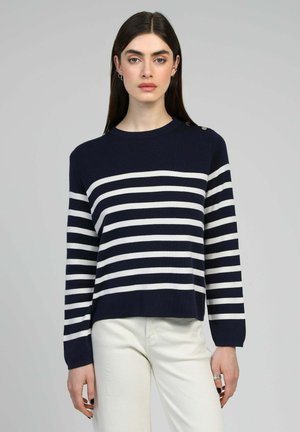 Femme portant un pull à rayures horizontales bleu marine et blanc à manches longues et un pantalon blanc, debout devant un fond gris uni.