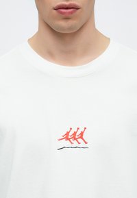 T-shirt en coton blanc avec un motif brodé rouge représentant trois figures sautant, au-dessus d'une signature noire, avec un col rond.