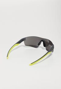 Lunettes de sport avec de grandes verres foncés, monture noire et embouts de tempes jaune vif, présentant un design incurvé et un matériel minimal.