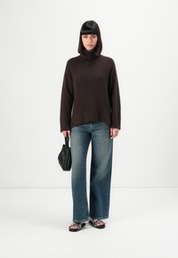 Femme portant un pull à col roulé marron foncé, un jean bleu ample, des sandales à imprimé serpent, tenant un petit sac à main noir sur fond blanc uni.