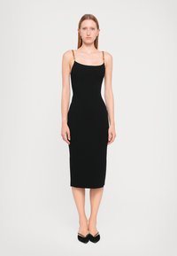Black fitted midi dress with ribbed texture, thin gold chain straps, and a square neckline. Paired with black pointed toe shoes.