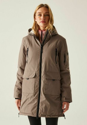 Donna che indossa un lungo cappotto invernale color tortora con cappuccio, cerniere nere e grandi tasche frontali, in piedi contro uno sfondo chiaro e uniforme.