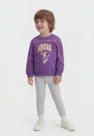 Ένα χαμογελαστό παιδί στέκεται φορώντας μωβ φούτερ Adidas με γραφικό της Minnie Mouse, ανοιχτό γκρι κολάν, λευκές κάλτσες και λευκά αθλητικά παπούτσια.