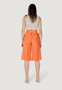 Culotte arancioni in lino con vita alta e dettaglio della cintura, abbinati a un crop top bianco in pizzo, che creano una silhouette rilassata.
