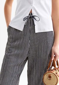 Top bianco con bottoni abbinato a pantaloni larghi a righe. Una persona tiene in mano una piccola borsa rotonda di vimini con manico.