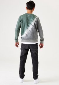 Felpa tie-dye verde e grigia con maniche lunghe e polsini a costine, abbinata a jeans in denim scuro e sneakers bianche.