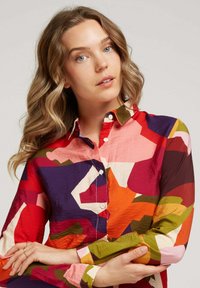 Chemise colorée avec un motif géométrique dans des nuances vibrantes de rouge, rose, violet, vert et orange ; fabriquée en tissu léger avec des boutons.
