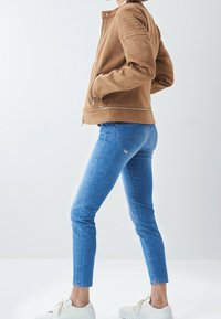 Hnědá kožená bunda s límečkem stojáčkem, prošívanými detaily na ramenou a stříbrnými zipy, ve spojení s modrými úzkými džíny a bílými teniskami.