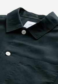 Chemise noire à manches courtes avec un col classique, deux boutons blancs et une poche poitrine. Texture lisse et design simple.