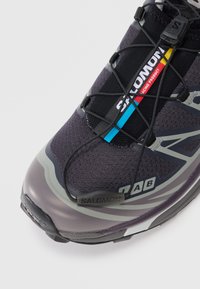 Schwarz-grauer Sportschuh mit strukturiertem Obermaterial, dünnen Schnürsenkeln, Gummikappe und Logoprägungen in Blau und Rot. Leichtes Design.