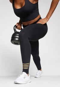 Črne športne pajkice z zlatimi črtami na gležnjih, v kombinaciji s črnimi kratkimi majicami. Vidne so bele športne čevlje in kettlebell.