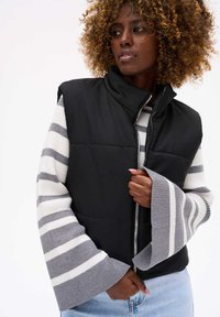 Czarna bezrękawnikowa kurtka puchowa z wysokim kołnierzem, w połączeniu z paskowanym szarym i białym swetrem z długim rękawem oraz jasnoniebieskimi dżinsami.