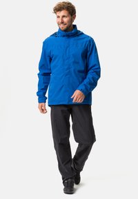 Blaue wasserdichte Jacke mit Kapuze, Reißverschluss und verstellbaren Bündchen, kombiniert mit schwarzen leichten Hosen und dunklen Schuhen.