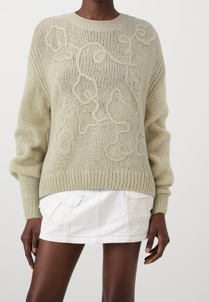 Beżowy sweter dziergany z wypukłym, abstrakcyjnym wzorem winorośli, zestawiony z białą mini spódnicą z przednimi kieszeniami naszywanymi.