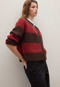 Cardigan rayé chunky en tricot rouge et marron. Présente une fermeture à boutons, des poignets côtelés et une silhouette courte. Assortie avec un pantalon noir.