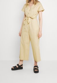 Gul mönstrad jumpsuit med korta ärmar, knappfront och bälte i midjan. Matchad med svarta klumpiga sandaler.