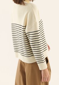 Suéter blanco acanalado con rayas negras horizontales, escote redondo y ajuste relajado. Combinado con pantalones marrones.