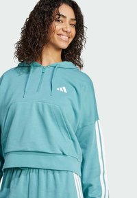 Turkoosi huppari, joka on valmistettu pehmeästä kankaasta. Siinä on vetoketjullinen kaula-aukko, kengurupussi ja valkoiset kolmiopuoliset koristeet hihoissa.