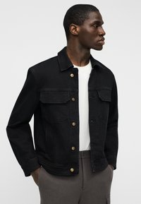 Μαύρο τζιν μπουφάν με κουμπιά μπροστά, δύο θήκες στο στήθος και κουμπιά σε χρυσό τόνο. Φορεμένο πάνω από λευκό T-shirt, παρουσιάζεται πάνω σε ουδέτερο φόντο.