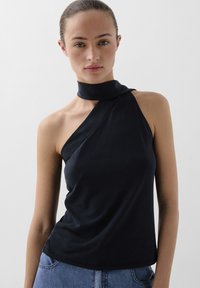 Zwarte mouwloze top met een hoge halslijn en een asymmetrisch wikkelontwerp. Zachte textuur en getailleerd silhouet, gecombineerd met casual blauwe spijkerbroek.