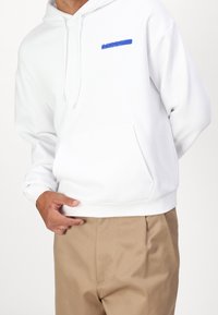 Sudadera blanca con capucha y bolsillo frontal, y logo azul de "Lacoste". El modelo lleva pantalones color beige. La tela parece suave y casual.