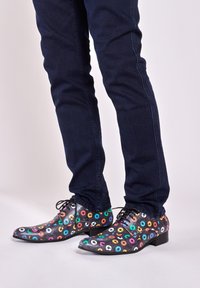 Zwarte schoenen met kleurrijke cirkelpatronen, voorzien van veters en een lichtpuntige neus, gecombineerd met donkere jeans.
