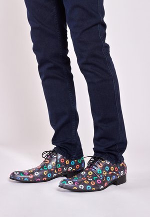 Zwarte schoenen met kleurrijke cirkelpatronen, voorzien van veters en een lichtpuntige neus, gecombineerd met donkere jeans.