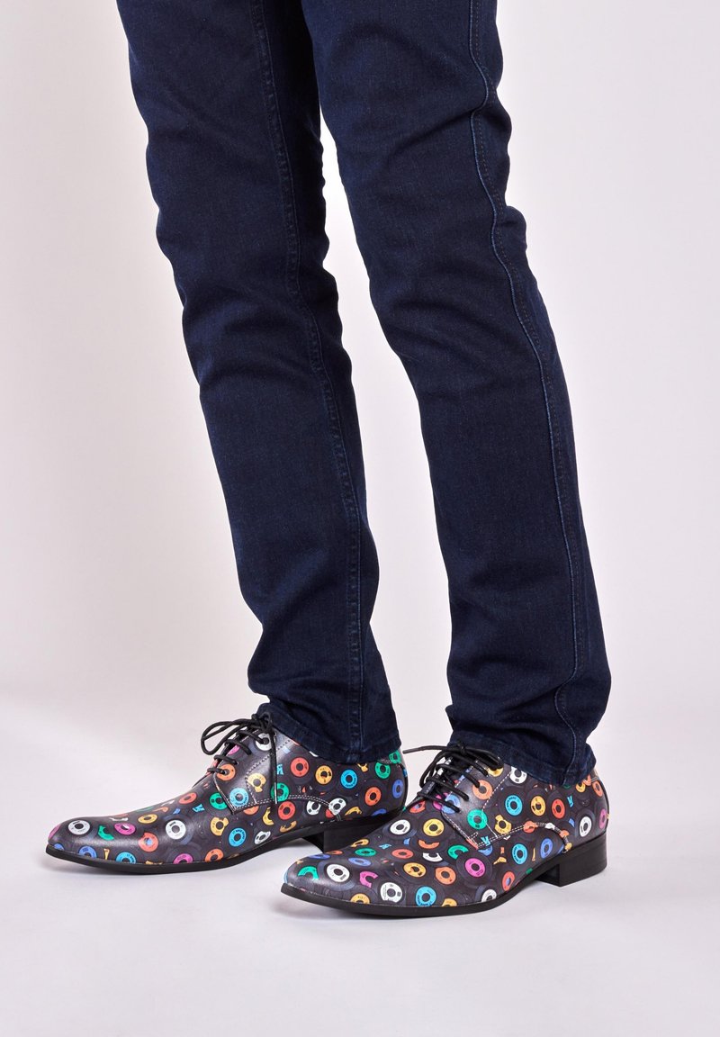Zwarte schoenen met kleurrijke cirkelpatronen, voorzien van veters en een lichtpuntige neus, gecombineerd met donkere jeans.