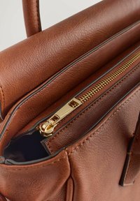 Hnědá kožená taška s hladkou texturou, vybavená zlatým zipem a vnitřní podšívkou. Design zahrnuje horní rukojeti a jemné šití.