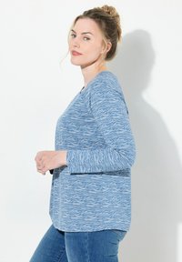 Top azul de manga larga con un patrón ondulado blanco, cuello redondo y aberturas laterales, combinado con jeans azules. Textura suave y corte relajado.