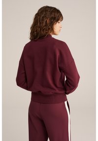 Felpa bordeaux con colletto alto, vita a coste e spalle scese. Abbinata a pantaloni coordinati con strisce bianche sui lati.