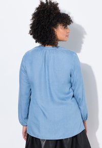 Camicia in denim azzurro chiaro con maniche lunghe, collo arricciato e orlo curvo. Texture morbida con dettagli di cucitura minimi.