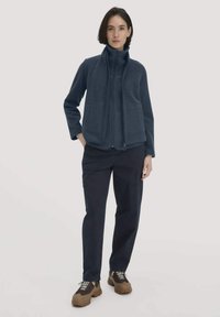 Vrouw staat, draagt een donkerblauwe fleecejack met rits, zwarte broek en bruine wandelschoenen tegen een effen lichte achtergrond.