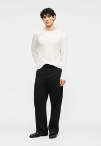 Άνδρας στέκεται με τα χέρια πίσω από την πλάτη, φοράει λευκό πουκάμισο μακρυμάνικο, μαύρο παντελόνι φαρδύ και μαύρα παπούτσια, σε απλό φόντο.