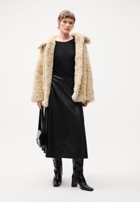 Faux fur crèmefarvet frakke, sort top, høj talje sort læder nederdel og knælange sorte støvler med hæl. Accessories inkluderer en sort håndtaske.