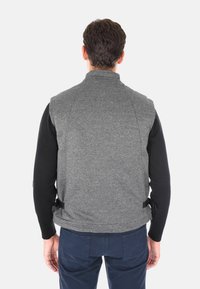 Gilet grigio con texture e colletto alto, con accenti neri e cinturini laterali, indossato sopra una maglietta nera a maniche lunghe.