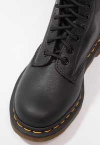 Bota de cuero negro con punta redondeada, diseño con cordones y costura amarilla a lo largo de la suela, con una superficie texturizada y herrajes negros.