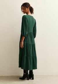 Maxikleid aus strukturiertem dunkelgrünem Stoff mit Puffärmeln und gestuftem Design, kombiniert mit knielangen schwarzen Stiefeln.