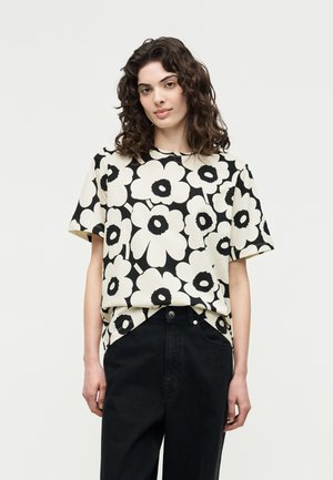 Mujer con cabello rizado que lleva una camisa de manga corta con estampado floral en blanco y negro y pantalones de cintura alta negros contra un fondo blanco.