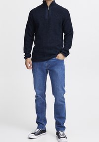 Marineblauer Reißverschluss-Pullover mit strukturiertem Strickdesign, kombiniert mit blauen Jeans und schwarzen Sneakers mit weißen Schnürsenkeln und Gummisohlen.