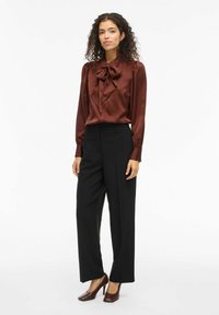 Brūna satīna blūze ar lielu loku apkakli, garām piedurknēm un pogu aizdari, apvienota ar augsta jostasvietas melnajām biksēm un brūnajiem papēžiem.